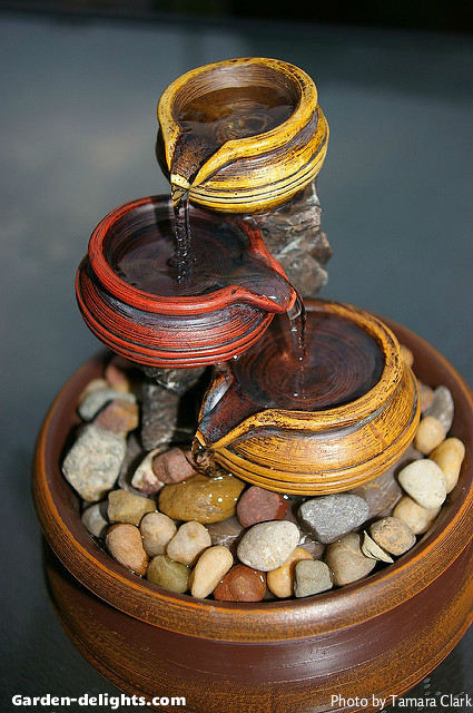 Small tabletop ceramic fountain with three small bowls pouring into each other that are yellow, red and orange with a rock base in a ceramic bowl that is a cordless fountain used for the patio table or inside your home or office, tabletop waterfalls, tiered tabletop fountains, fiberglass fountains, indoor water fountains, indoor tabletop water feature, tabletop Gardens, mini fountains, fountain ideas, desktop fountains create a visual motion and sensory sound of water allowing for a unique visual artistic theme.
