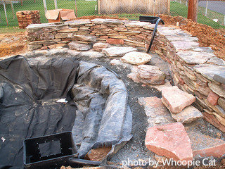 Flexible pond liner inside of dugout pond with shale stone stacked up like a wall along edge. Fecorative pond ideas, backyard water fe ature planning.