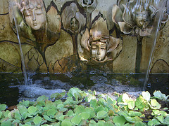 Mystical fairy faces on the front of wall fountain pouring water into lower basin with plants, ferry water fountains, fantasy fairy fountains.