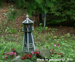Small miniature green lighthouse beside rocks and red flowers with Windows in beacon room, outdoor lighthouses, nautical garden decor.